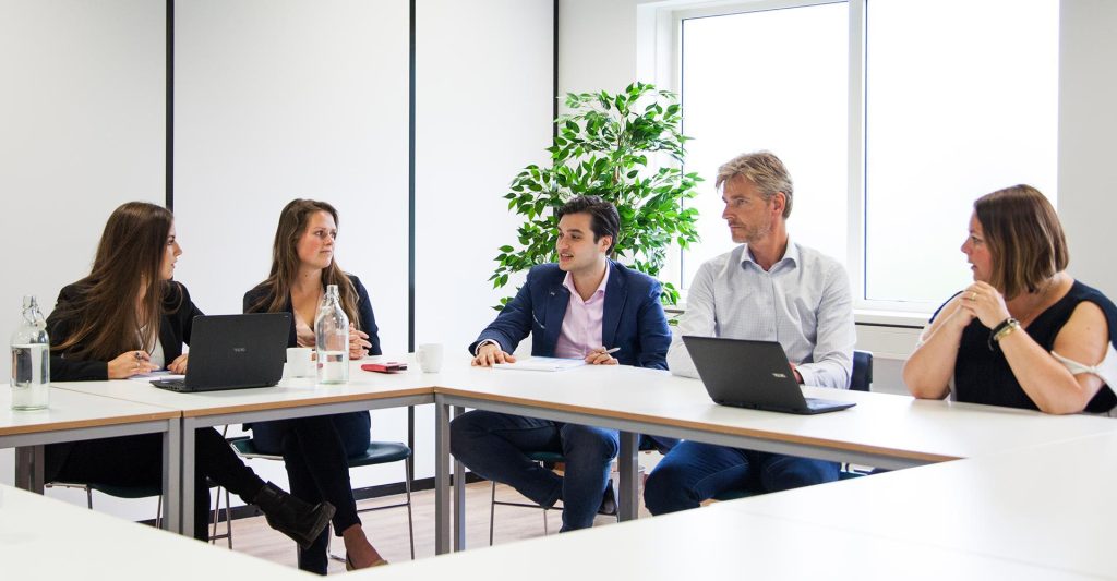 Teamvergadering aan tafel met laptops en notitieblokken.