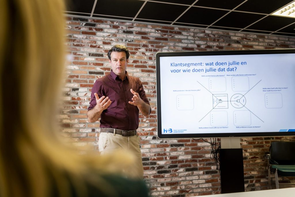 Man geeft presentatie voor publiek, scherm toont tekst.