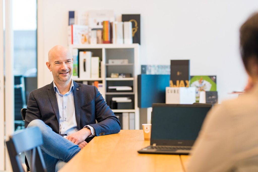 Man in een vergaderruimte met laptop en boekenplank.