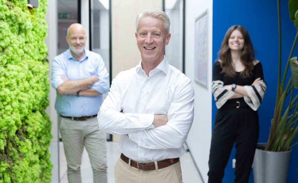 Drie mensen poseren lachend in kantoor.