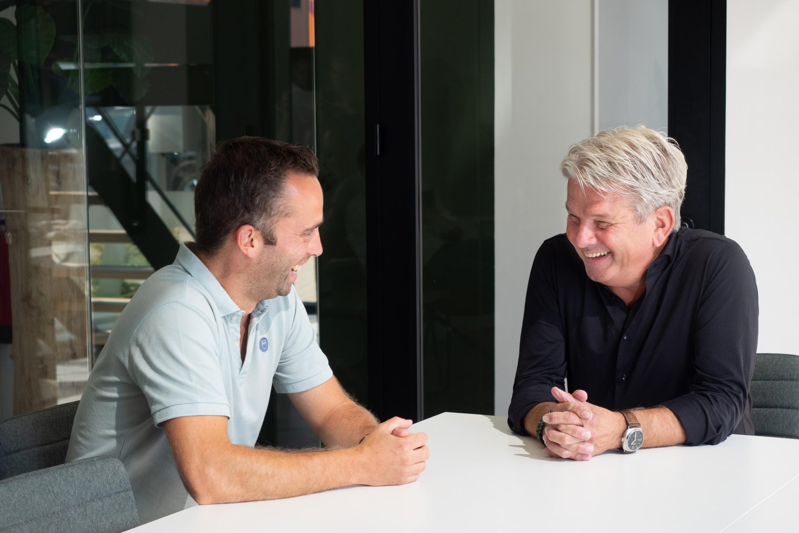 Twee lachende mannen praten aan een tafel.