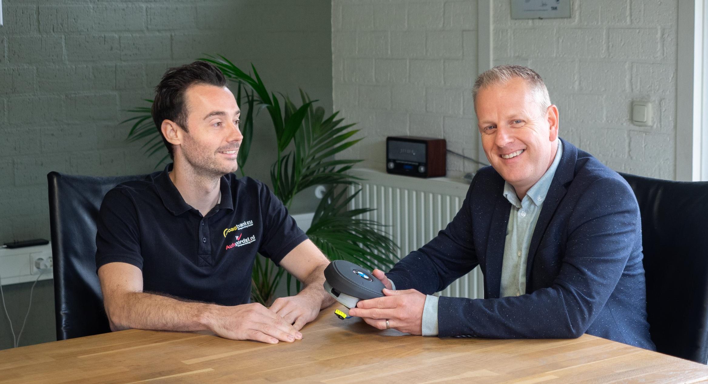 Twee mannen met auto-onderdeel aan tafel.