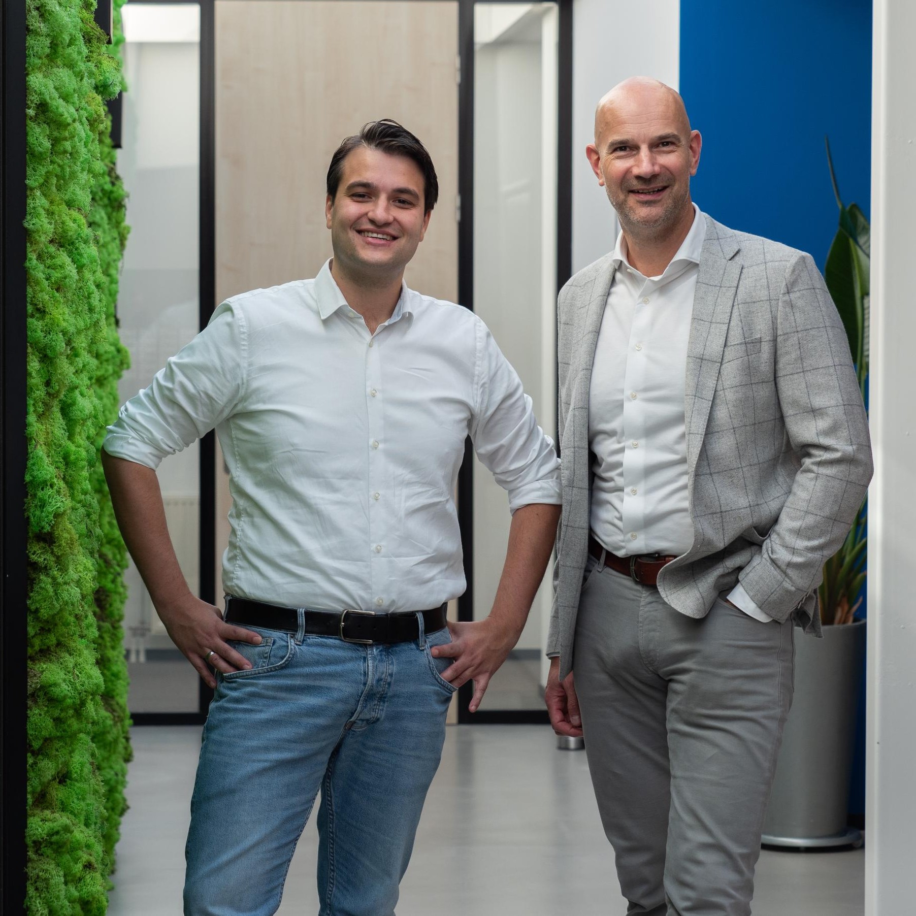 Twee mannen poseren in een moderne kantooromgeving.