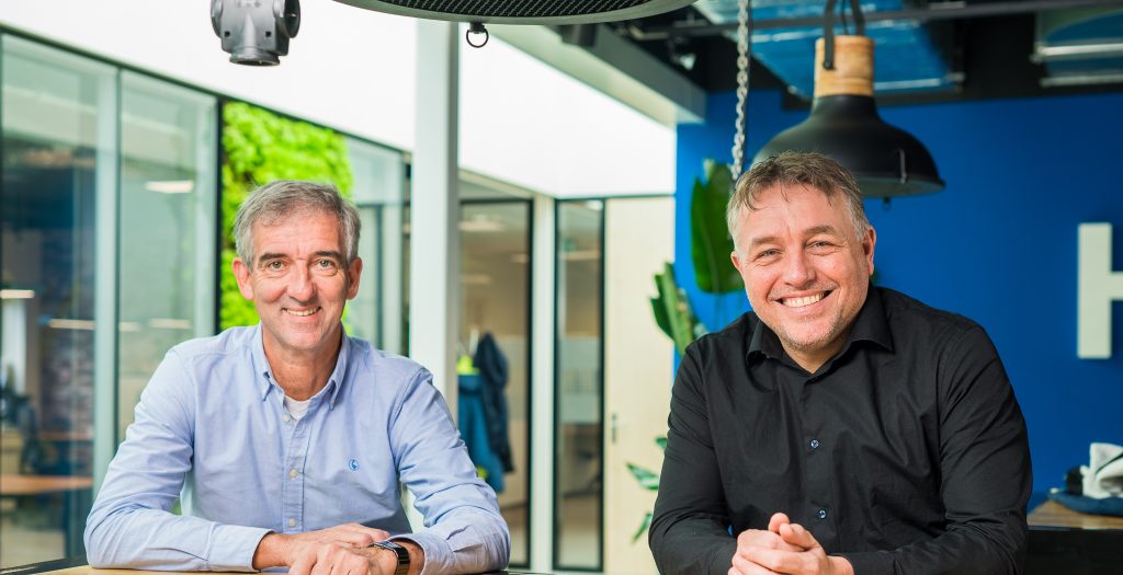 Twee mannen glimlachen zittend aan tafel in kantoor.