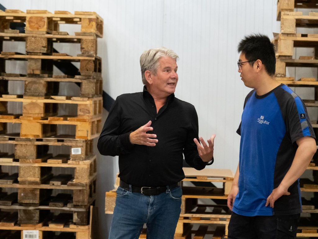 Twee mannen praten voor stapel pallets in magazijn.